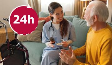 Eine Pflegerin reicht einem alten Mann Medikamente, die beiden sitzen ein einem gemütlichen Wohnzimmer