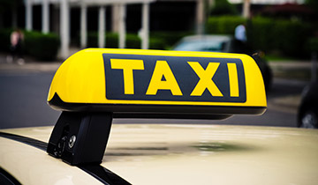Gelbes Taxi-Dachschild mit schwarzer Aufschrift „TAXI“ auf einem Fahrzeug, im Hintergrund unscharfe Straße mit Gebäuden und Passanten