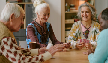 Ältere Menschen sitzen gemeinsam an einem Tisch und spielen Karten, lachen und unterhalten sich – eine fröhliche Freizeitaktivität für Senioren zu Hause