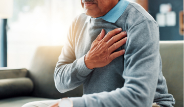 Älterer Mann sitzt auf einem Sofa und hält sich die schmerzende Brust, was ein mögliches Anzeichen für Herzinfarkt ist.