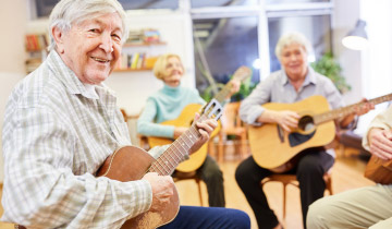 Senioren sitzen gemeinsam in einem Raum und spielen Gitarre, ein Senior im Vordergrund lächelt in die Kamera