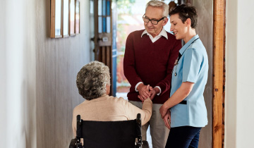 Pflegebedürftige Frau im Rollstuhl wird in einem hellen Flur von ihrem gesunden Ehemann und einer Pflegerin besucht und begrüßt