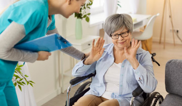 Pflegekraft spricht mit einer Seniorin im Rollstuhl, die durch eine abwehrende Handbewegung ihre Pflegeverweigerung ausdrückt