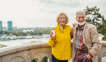 Eine Rentnerin und ein Rentner stehen Arm in Arm an einer Brücke und sehen glücklich aus, sie trinkt ein Heißgetränk in einem Togo-Becher