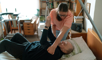 Eine Frau als pflegende Angeh�rige unterst�tzt bettl�gerigen Senior beim Aufrichten im h�uslichen Pflegeumfeld.