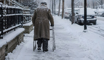 Ältere Person von hinten mit Rollator auf einem verschneiten Gehweg im Winter