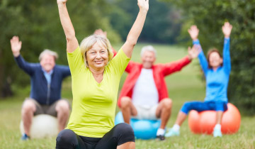 Eine Gruppe älterer, fröhlicher Personen in leichter Sportkleidung sitzt im Freien jeder auf einem Gymnastikball und recken die Arme in die Höhe. Eine Frau im Vordergrund ist scharf im Bild, drei weitere Personen im Hintergrund sind unscharf