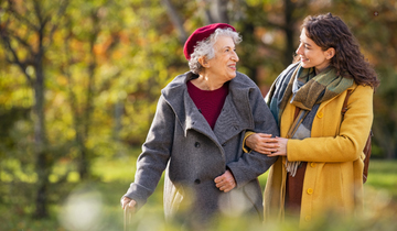 Seniorin und junge Frau laufen lächelnd Arm in Arm durch einen Park.