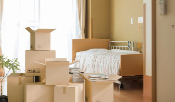 Umzugskartons und verpackte Gegenstände in einem hellen Schlafzimmer mit Bett, vorbereitet für den Umzug ins Seniorenwohnheim