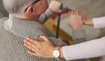 Pflegekraft unterstützt einen älteren Menschen beim Aufstehen und legt eine Hand auf dessen Schulter, symbolisches Bild für Unterstützung im Alter beim betreuten Wohnen
