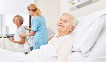 Ältere, bettlägerige Patientin liegt lächelnd im Pflegebett, während Pflegekräfte im Hintergrund eine weitere Patientin versorgen