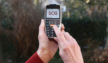 Eine Hand hält ein Seniorentelefon. Der Finger der anderen drückt eine Taste. Auf dem Display steht „SOS“.