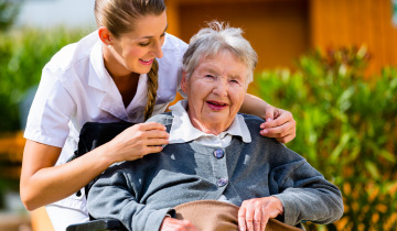 Eine Pflegerin beugt sich freundlich über eine alte Dame im Rollstuhl