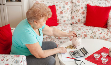 Ältere Frau sitzend auf dem Sofa. Auf dem Tisch vor ihr liegen ein Taschenrechner und Münzen, die sie in das ebenfalls auf dem Tisch stehende Sparschwein steckt