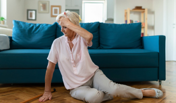 Eine ältere Dame sitzt am Boden vor ihrem Sofa und hält sich den Kopf, weil sie gestürzt ist