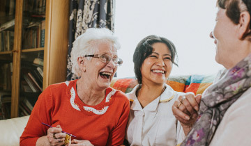 Drei Frauen lachen fröhlich miteinander, zwei sind Seniorinnen, eine ist eine junge Begleitperson