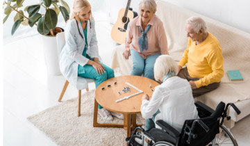 Eine Gruppe Senioren sitzt am Tisch und spielt Domino, während eine Pflegerin zuschaut