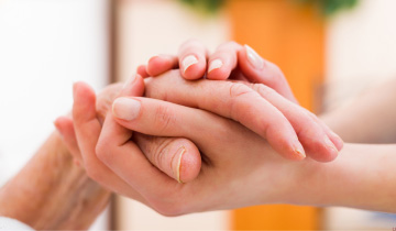Die Hand einer älteren, pflegebedürftigen Frau liegt in den umsorgenden Händen einer jungen Frau