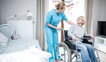 Eine weiblich Pflegekraft hilft einer älteren Dame, die im Rollstuhl sitzt, in ihrem Schlafzimmer