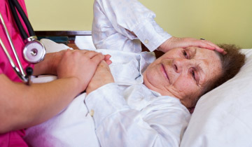 Eine bettlägerige, ältere Dame lächelt ihre Pflegekraft an, die der Patientin die Hand hält