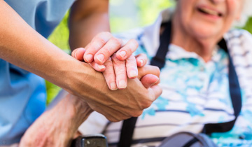 Im Vordergrund ergreift eine ältere Hand die helfende Hand einer Pflegeperson, im Hintergrund sieht man die ältere Person mit viel Tiefenunschärfe
