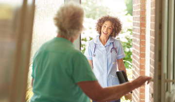 Eine Pflegekraft steht vor der Haustür und wird von einer älteren Dame hereingelassen