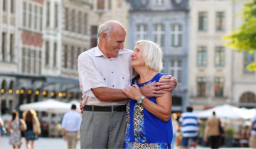 Ein älteres Pärchen steht Arm in Arm auf einem Marktplatz mit Häuserfassaden im Hintergrund und schaut sich glücklich an, als Sinnbild für die seniorenfreundliche Stadt