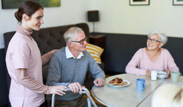 Pflegekraft unterstützt einen älteren Mann mit Rollator, während er gemeinsam mit einer älteren Frau an einem Tisch sitzt und Kaffee in einem Seniorenheim oder einer Seniorenresidenz trinkt