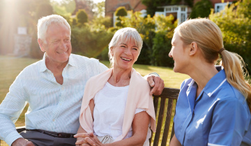 Zwei Senioren sitzen auf einer Bank an einem sonnigen Tag und lächeln eine Pflegefachkraft an, die ebenfalls auf der Bank sitzt.